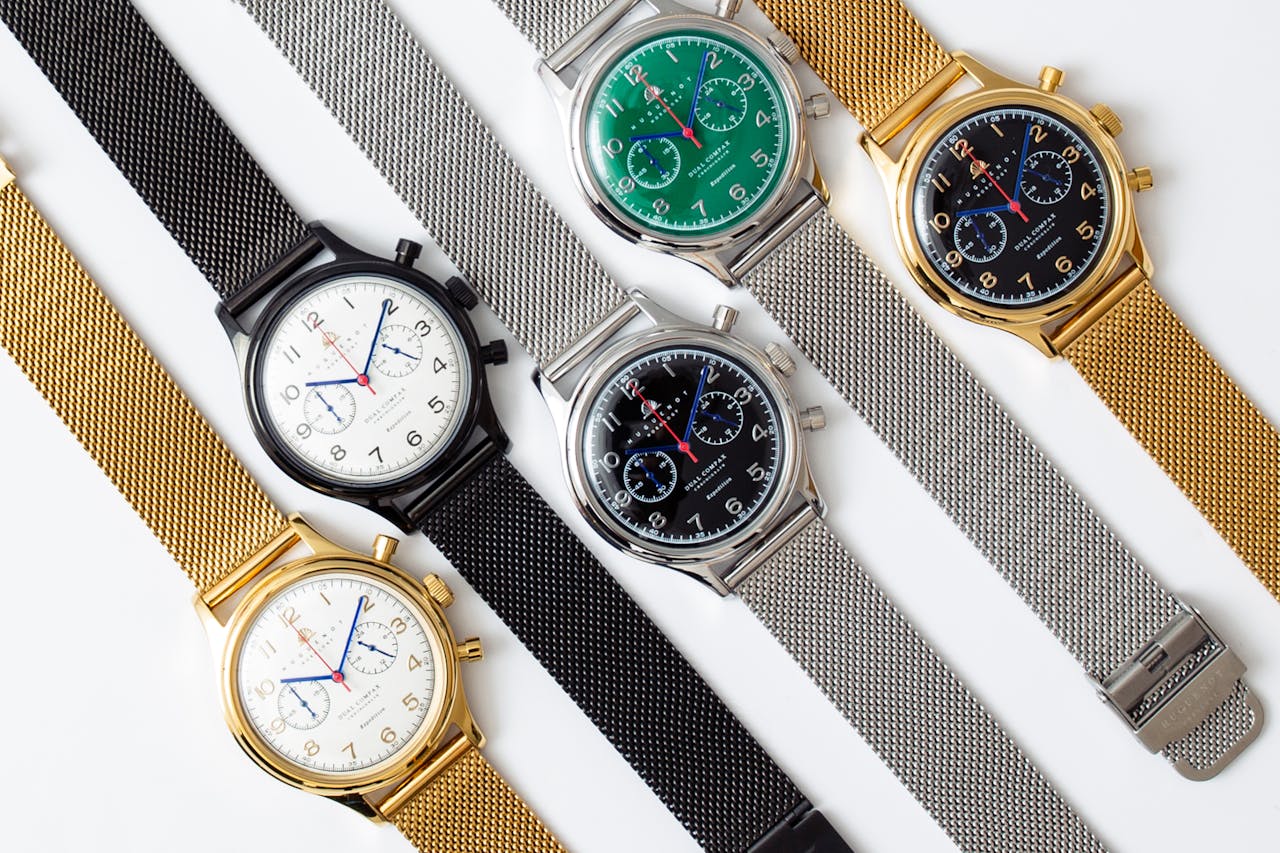 A display of five analog wristwatches with various colored dials and metal straps on a white background.